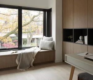 Modern minimalist window seat with light oak cabinetry, neutral cushions, throw blanket, and built-in shelving next to a light wood desk.