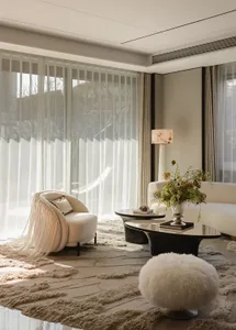 Bright modern luxury living room featuring a cream boucle fringed armchair, white fluffy ottoman, dark marble coffee table, and layered neutral rugs with large windows letting in natural light.
