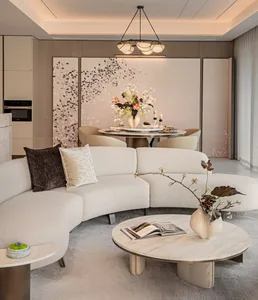 Modern minimalist living and dining room featuring a large curved bouclé sectional sofa, round travertine coffee table, and a dining set against a floral accent wall with a sculptural chandelier overhead.