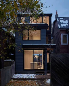 Modern dark grey house with large illuminated windows showing warm wooden interior and a dog standing in the living area during winter dusk.