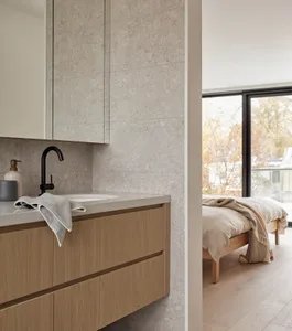 Modern bathroom featuring a travertine tiled wall and countertop, light oak floating vanity with black matte faucet, and a view into a minimalist bedroom with wooden bed and large window.