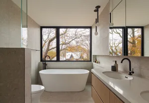 Modern minimalist bathroom with travertine tile walls, white freestanding bathtub, wooden vanity with black faucet, large black-framed windows showing autumn trees, and a white toilet.