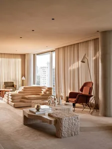 Modern organic living room featuring a large beige modular sofa, travertine coffee tables, a textured terracotta armchair, and floor-to-ceiling sheer curtains with city view.