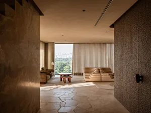 Modern living room featuring textured travertine walls and floor, a large beige modular sofa, wooden coffee table, and large windows revealing a green city skyline.