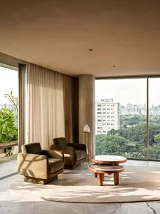 Modern living room with two olive-green velvet armchairs, round wooden coffee table, large curved beige rug, and floor-to-ceiling windows overlooking a city skyline and lush greenery.