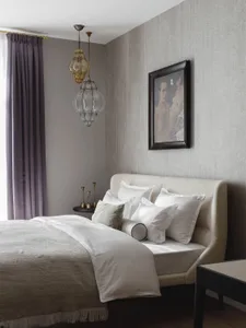 Modern classic bedroom featuring a beige upholstered bed with multiple white pillows, textured grey walls, two ornate glass pendant lights, purple curtains, and a framed painting of figures.