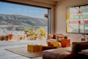 Bright modern living room with large floor-to-ceiling windows overlooking mountains, filled with potted plants in terracotta pots, yellow and orange furniture, and a brown leather sofa.
