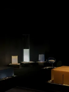Collection of modern sculptural table lamps made of glass and illuminated from within, displayed on dark pedestals in a minimalist gallery setting.