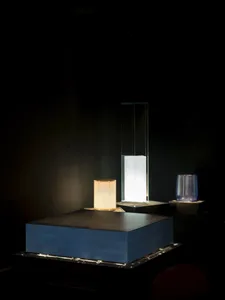 Three contemporary glass table lamps of varying heights and shapes glowing warmly on a large blue stone platform in a dark exhibition space.