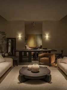 Modern minimalist living and dining area featuring a large round dark coffee table with textured ceramic vases, a mustard bouclé armchair, cream bouclé sofas, warm wall sconces, and abstract art in the background.