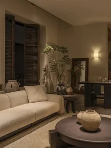 Modern organic living room featuring a cream boucle sofa, dark wood shutters, multiple ceramic vases, a round coffee table, and warm wall lighting.