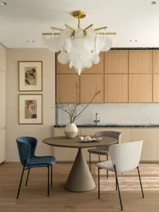 Modern dining area featuring a round concrete pedestal table, one blue velvet chair and two bouclé upholstered chairs, set against a light oak kitchen cabinet wall with a large artistic chandelier.