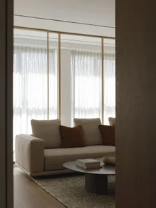 Modern minimalist living room featuring a beige boucle sofa with brown velvet cushions, round coffee table, and sheer curtains allowing diffused daylight.