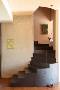 Modern brutalist interior featuring a dark concrete floating staircase, raw concrete pillar, warm wooden floor, and two colorful abstract paintings with a red pendant lamp.