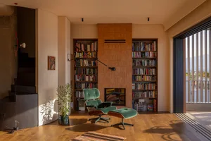 Mid-century modern living room with built-in teak bookshelf wall, emerald green velvet Eames-style lounge chair and ottoman, wooden floor, potted plant, and large glass door leading to a balcony with mountain view.