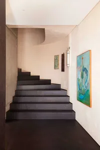 Modern interior view of a dark floating staircase with curved micro-cement walls, dark floor, and colorful abstract paintings on a light beige wall.