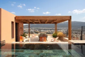Modern rooftop terrace with infinity pool, wooden pergola, dining table, lounge chairs, and panoramic mountain and city views under a clear blue sky.