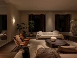Modern organic living room featuring a creamy white boucle sofa, two brown wooden lounge chairs, a large round dark wood coffee table, textured area rug, and multiple potted plants against a neutral backdrop with wooden shutters.