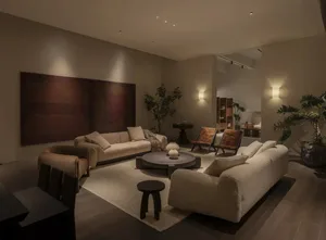 Modern living room featuring two boucle sofas, round coffee table, textured burgundy artwork, wooden accent chairs, and multiple indoor plants under warm ambient lighting.