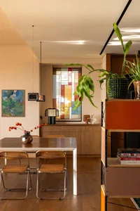 Modern dining room featuring a walnut sideboard, cane-backed folding chairs, a large stained-glass window, and warm neutral walls with wooden flooring.
