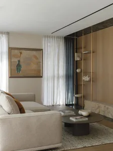 Modern living room featuring a beige sectional sofa, round dark coffee table on a textured rug, tall brass and glass shelving unit against a wood-paneled wall with marble base, and an abstract painting on the opposite wall.