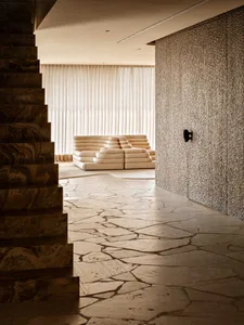 Modern minimalist living room featuring a large cream modular sofa, irregular travertine stone floor, dark marble staircase, and textured beige wall panel.