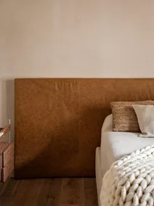 Close-up of a large brown leather upholstered headboard with visible texture, paired with cream knit bedding, a chunky white knitted throw, and a wooden nightstand on a dark wood floor.