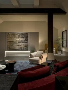 Modern luxury living room featuring a deep red velvet sofa in the foreground, a cream swivel armchair, large abstract painting with backlighting, and blue textured carpet.
