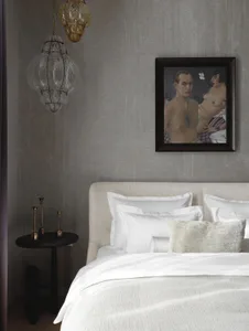 Modern bedroom with textured grey walls, white linen bedding, black side table with brass candlesticks, hanging glass lanterns, and a framed painting of a semi-nude couple.