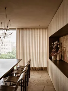 Modern minimalist dining area featuring a dark breakfast bar, four brown leather bar stools, irregular travertine stone floor, sheer beige curtains, wooden cabinetry, and a sculptural glass pendant light.