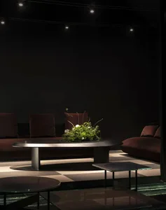Dark moody living room featuring deep burgundy velvet sofas, a large oval stone coffee table with an elaborate green and white floral arrangement, checkered floor, and dramatic ceiling track lighting.