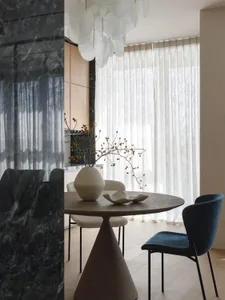 Modern minimalist dining area featuring a round dark marble pedestal table, blue velvet dining chair, light wood cabinetry, and a dramatic white petal chandelier.