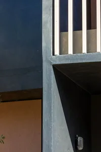 Close-up architectural detail of a raw concrete corner where deep blue, terracotta, and black painted surfaces meet under bright sunlight, creating sharp shadows.