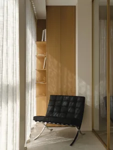 A black Barcelona chair placed in a bright minimalist reading nook with built-in oak shelving and sheer white curtains.