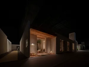 Night view of a modern exhibition booth inside a large exhibition hall, featuring a prominent reclaimed wood beam over the entrance, warm lighting, stone accents, and a glimpse of a contemporary living area with wooden furniture and plants.