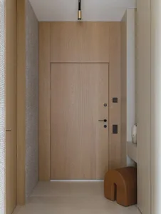 Modern minimalist entryway with light oak wood wall paneling, a flush wooden door, black hardware, a curved brown leather stool, and a narrow console shelf.