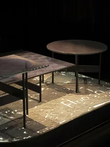 Two modern coffee tables, one rectangular with a glass top revealing a mosaic pattern and another round solid bronze top, placed on a thick textured rug with projected light patterns in a dark room.