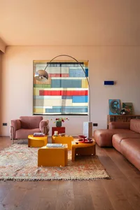 Bright living room with peach walls, a pink velvet armchair, brown leather sofa, glossy yellow nesting tables, a large colorful geometric tapestry, and a patterned area rug on wooden floors.
