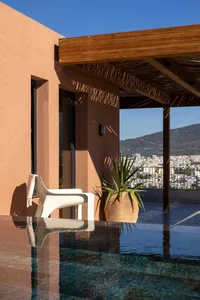 Modern rooftop terrace featuring terracotta plaster walls, wooden pergola, white lounge chair, potted agave plant, and a reflective infinity pool with mountain and city views.