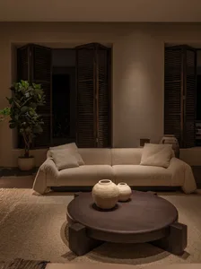 Modern living room with a cream boucle sofa, round dark wood coffee table, textured ceramic vases, potted fiddle leaf fig, and dark wooden louvered shutters.