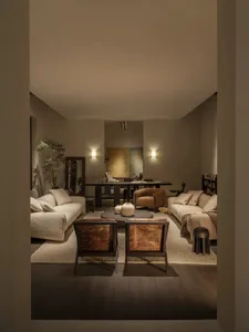 View into a modern open-plan living room featuring two white boucle sofas, two dark wood cowhide accent chairs, a round coffee table with ceramic vases, warm wall sconces, and a dining area in the background.