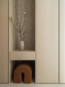 Modern minimalist entryway featuring a built-in beige niche with a crackle-glaze ceramic vase holding flowering branches, set against vertically textured neutral wallpaper, with a brown leather arched stool tucked underneath.