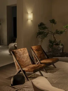 Two brown cowhide lounge chairs with dark wood frames placed on a textured rug in a warmly lit modern living room corner, accompanied by a large potted plant and wall sconce.