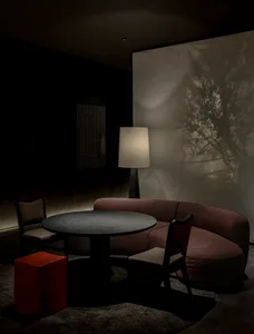 Modern moody dining area featuring a curved burgundy textured sofa, round dark wood table, matching chairs, a bright red cube stool, and dramatic tree shadow projection on the wall.