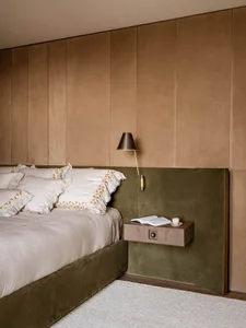 Modern bedroom featuring a sage green velvet bed base and headboard, layered with cream linen bedding with geometric embroidery, set against a two-tone wall with taupe suede upper panels and deep green velvet lower paneling, accompanied by a floating walnut nightstand and brass wall lamp.