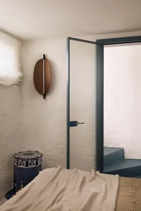 Modern bedroom corner with off-white textured plaster walls, dark blue metal-framed ribbed glass door, walnut oval wall lamp, and ornate indigo hexagonal side table.