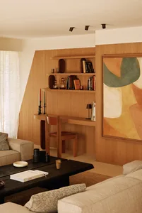 Modern living room featuring a built-in oak desk and shelving unit, large abstract painting, dark coffee table, and beige sofa with natural daylight.