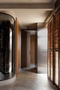 Modern minimalist hallway featuring a large curved walnut wooden pivot door, wooden shutters, concrete floor, and warm natural light.