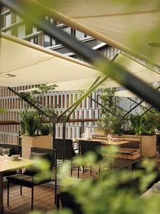 Modern rooftop terrace restaurant with teak dining tables, dark woven chairs, large retractable fabric umbrellas, and abundant potted plants against a contemporary building facade.