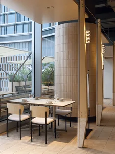 Modern restaurant interior featuring a curved light-wood slat column, minimalist dining table with light wood chairs, and large windows overlooking an outdoor patio with umbrellas.